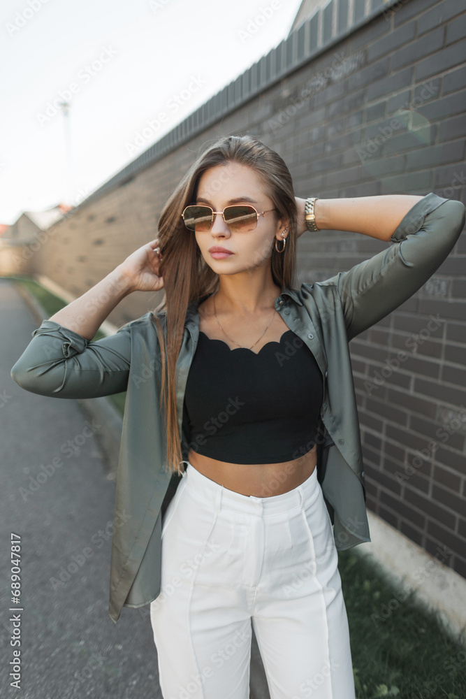 Fashion beautiful young stylish girl model with vintage sunglasses in fashionable clothes with a shirt standing near a black brick wall on the street