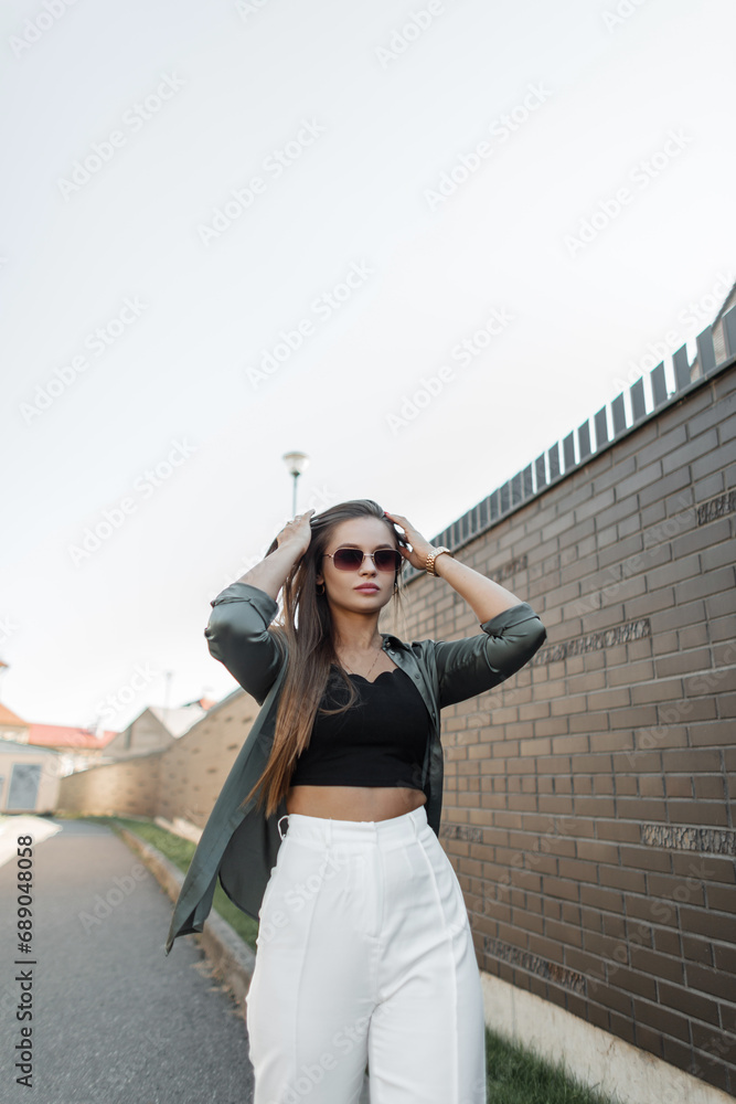Beautiful fashionable street girl model with sunglasses in a fashion outfit with a shirt walks in the city near a black brick wall