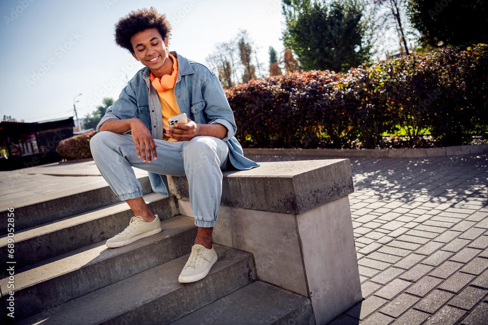 Portrait of intelligent friendly guy with chevelure wear denim shirt jeans headphones sit read notification on smartpone in park outdoors Portrait of intelligent friendly guy with chevelure wear denim shirt jeans headphones sit read notification on smartpone in park outdoors