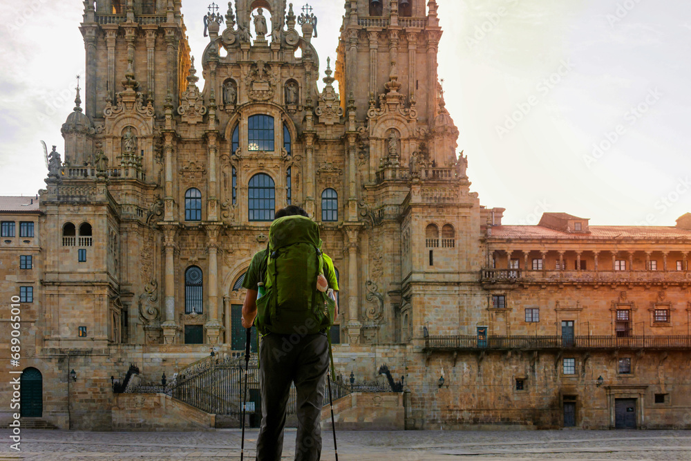 Traveler on the Camino de Santiago, with a backpack decorated with a scallop shell and two walking sticks in his hand, standing in the square of the Cathedral of Santiago de Compostela