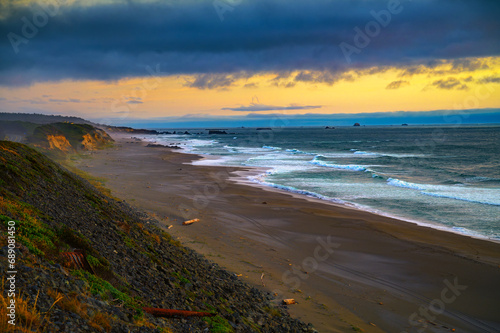 A serene sunset at a beach near Gold Beach, Oregon, showcasing dramatic cliffs under a canopy of heavy clouds, bathed in stunning yellow and orange evening light.