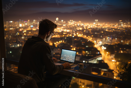 A male freelancer working from home on a laptop near a window in a stylish apartment overlooking the evening metropolis.