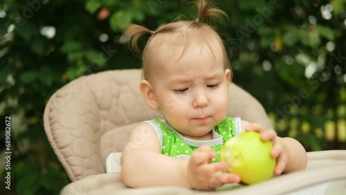 Portrait of a cute funny baby girl, eating a green apple, sitting in a high chair in the garden on a sunny summer day.