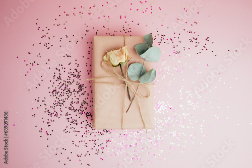 Fototapeta Naklejka Na Ścianę i Meble -  Background for greetings. Gifts wrapping in soft pink paper with dry eucalyptus branch on a light pink background with sequins in the shape of a heart. Top view