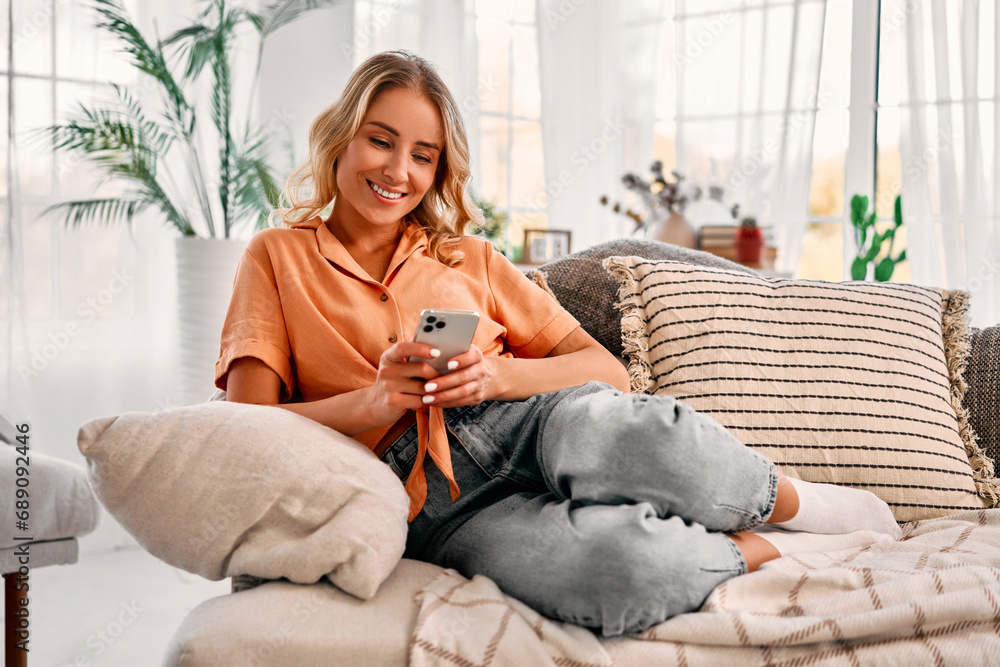 Leisure with gadgets. Relaxed caucasian woman in casual attire sitting on comfy couch with modern smartphone in hands. Smiling lady with blond hair scrolling social media during free time at home. Leisure with gadgets. Relaxed caucasian woman in casual attire sitting on comfy couch with modern smartphone in hands. Smiling lady with blond hair scrolling social media during free time at home.