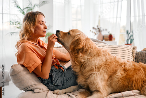 Domestic activities. Friendly golden retriever reaching out muzzle to attractive adult woman sitting on sofa at living room. Positive young blonde playing with favorite furry pet during pastime.