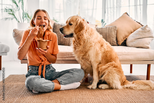 Family lifestyles. Happy blonde enjoying chinese noodles while golden retriever sitting near an expressing interest in delivered food. Furry adult dog asking for human meal at bright living room. photo