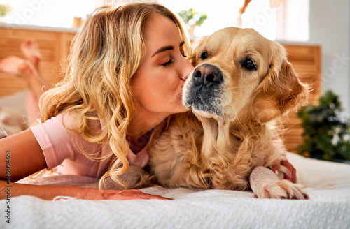 Sincere love to pet. Friendly young woman kissing muzzle of golden retriever while lying together on soft bed. Female blonde strengthening friendship bond with canine buddy at home.