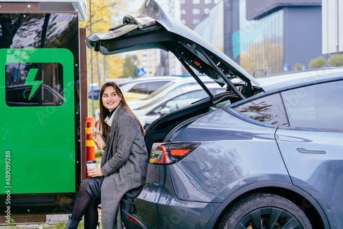 Young woman waiting for her electric car charging, sitting in a car trunk and drinking coffee. Alternative energy and rechargeable electric vehicle for sustainable eco friendly travel.