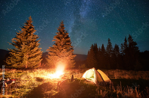 Night camping in mountains under starry sky and Milky way. Female tourist resting near burning campfire and illuminated tent in campsite, lying on grass and enjoying night sky with stars.