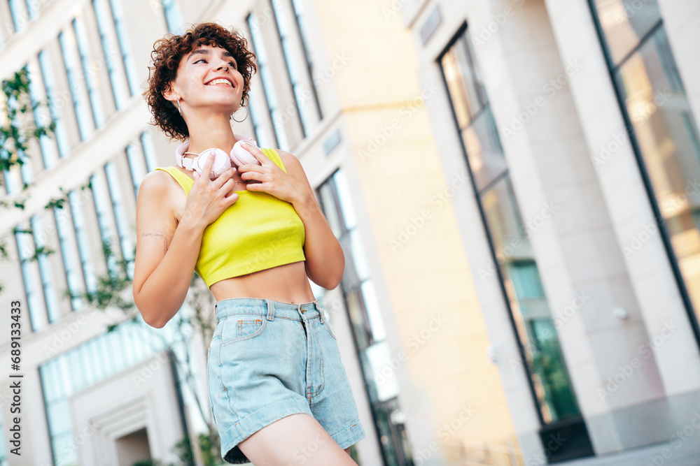 Young beautiful smiling hipster woman in trendy summer clothes. Carefree woman with curls hairstyle, posing in the street at sunny day. Positive model outdoors. Listens music at her headphones on neck Young beautiful smiling hipster woman in trendy summer clothes. Carefree woman with curls hairstyle, posing in the street at sunny day. Positive model outdoors. Listens music at her headphones on neck