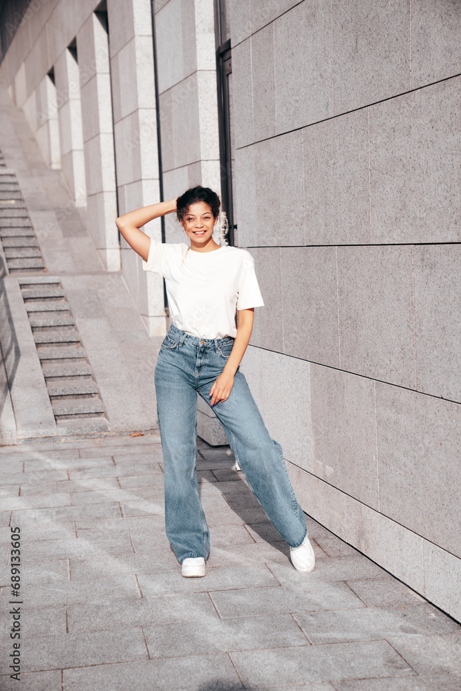 Young beautiful smiling hipster woman in trendy summer white t-shirt and jeans clothes. Carefree woman, posing in the street at sunny day. Positive model outdoors near wall Young beautiful smiling hipster woman in trendy summer white t-shirt and jeans clothes. Carefree woman, posing in the street at sunny day. Positive model outdoors near wall