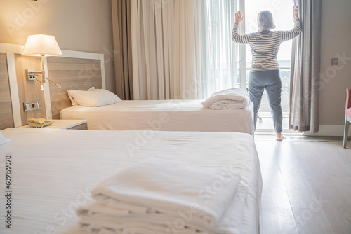 Fototapeta Naklejka Na Ścianę i Meble -  woman traveler opening the curtains and looking at the view from the window of a hotel room while on summer vacation travel concept