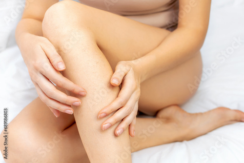 Young Caucasian woman touches her bare hairy legs with her hands. The female sits at home on a white bed. Hair removal, depilation procedures for beauty. Naturalness and body care concept.
