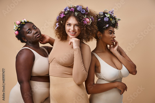 joyful multiethnic girlfriends in lingerie with colorful flowers in hair on beige, plus size beauty