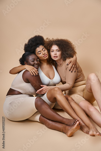 multiethnic plus size female friends in underwear sitting and embracing on grey, togetherness © LIGHTFIELD STUDIOS