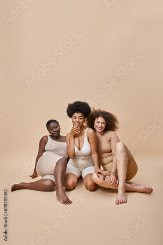 three joyful multiracial plus size women in underwear sitting on beige, natural body positive beauty
