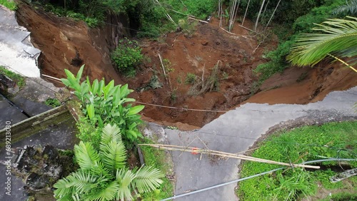 Camera fly up and show landslide area from above, village road destroyed, ground from steep slope of ravine collapsed down. House fence hang over cliff, destabilized soil is prone to further collapse photo