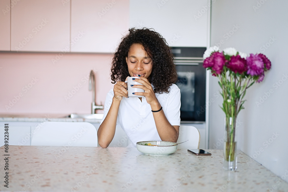 Beautiful young woman sitting at kitchen counter having breakfast and typing a text message using smart phone, relaxing at home in the morning     