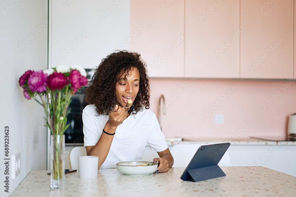 Beautiful young woman sitting at kitchen counter having breakfast and using tablet, relaxing at home in the morning. Working with computer in the office at home Beautiful young woman sitting at kitchen counter having breakfast and using tablet, relaxing at home in the morning. Working with computer in the office at home