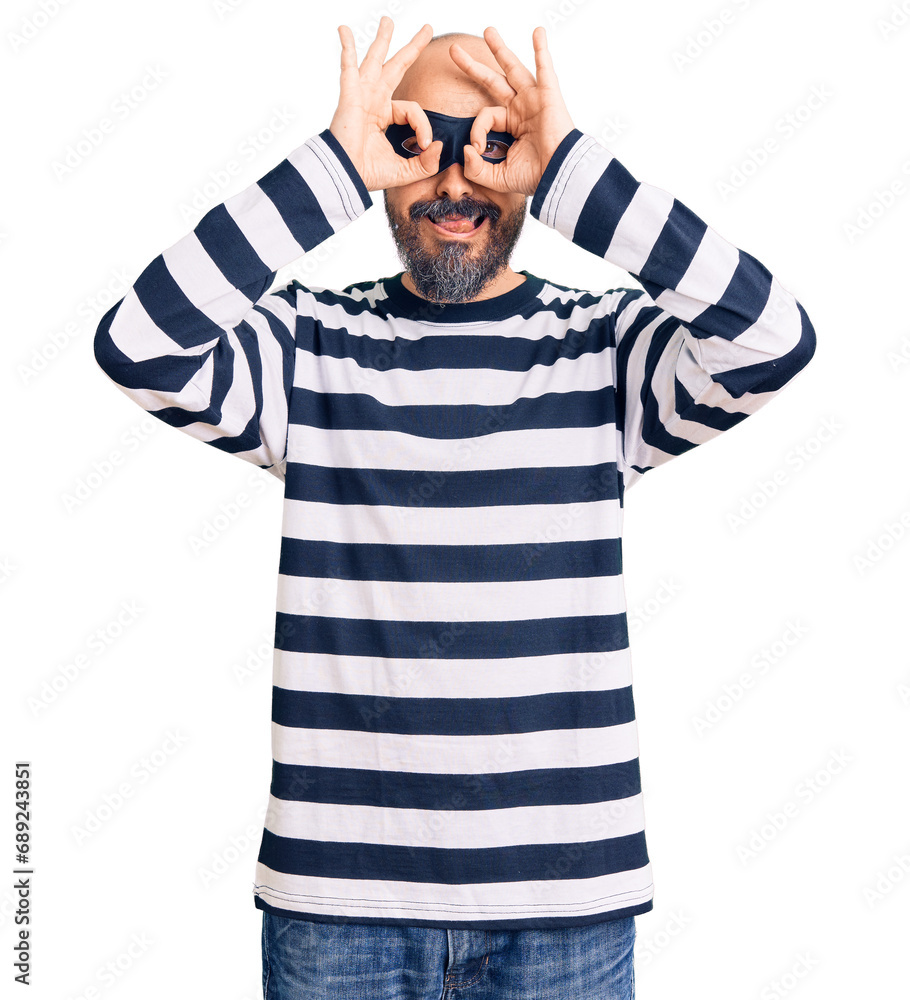 Young handsome man wearing burglar mask doing ok gesture like binoculars sticking tongue out, eyes looking through fingers. crazy expression. Young handsome man wearing burglar mask doing ok gesture like binoculars sticking tongue out, eyes looking through fingers. crazy expression.