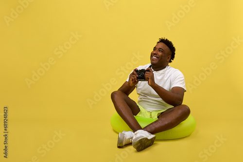 Fototapeta Naklejka Na Ścianę i Meble -  Positive young African man in a white T-shirt sits in a lifebuoy holding a camera. Cheerful photographer, a man in a funny pose, placed in a swimming circle on a yellow background