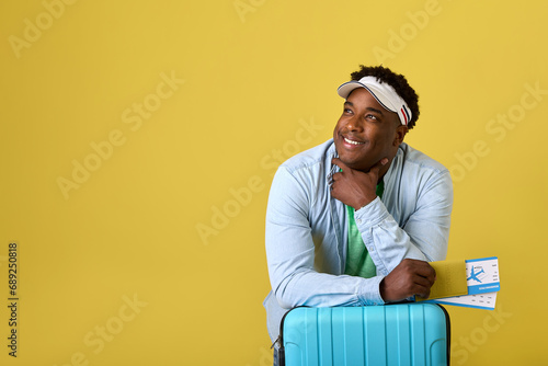 On yellow background African man in denim shirt and cap holds plane tickets in hand, looking into distance. A black man is traveler with blue suitcase waiting for announcement of boarding a flight © Aleksandr