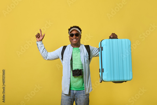 Happy young African man in sunglasses holding a large blue suitcase in his hand, pointing his finger up. Dark-skinned male traveler model demonstrates a roomy suitcase for trips on a yellow background