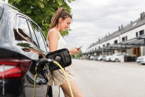 Young woman, putting a charger in an electric car parked in the parking area and adjusting an EV charging app on a mobile phone © 24K-Production