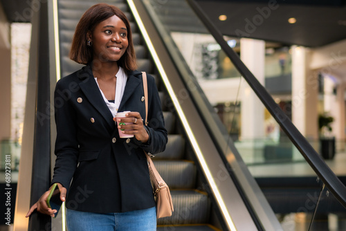In a joyful mood, the formally dressed black business woman holds her coffee and phone while descending on the escalator. photo