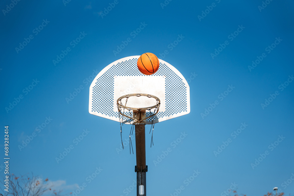 Basketball basket seen from below and from the front while an orange ball is falling from above. Contrasted with a blue sky on a sunny, clear day. Basketball basket seen from below and from the front while an orange ball is falling from above. Contrasted with a blue sky on a sunny, clear day.