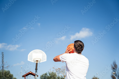 Young Caucasian boy with his back turned, wearing a white t-shirt throwing a basketball at a metal basket and the clear blue sky on a sunny day. photo