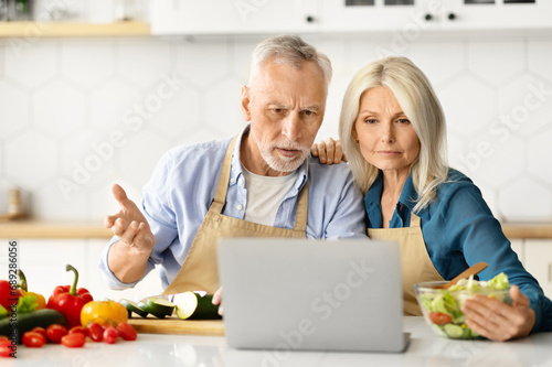 Wrong Recipe. Confused Senior Spouses Looking At Laptop While Cooking In Kitchen