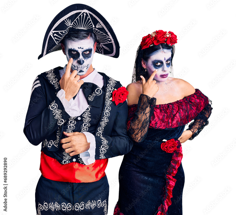 Young couple wearing mexican day of the dead costume over background pointing to the eye watching you gesture, suspicious expression