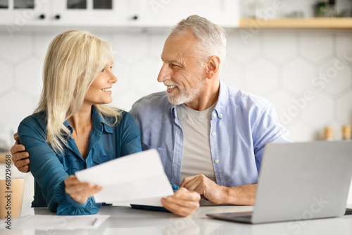 Portrait Of Cheerful Elderly Spouses Sitting In Kitchen And Checking Financial Papers