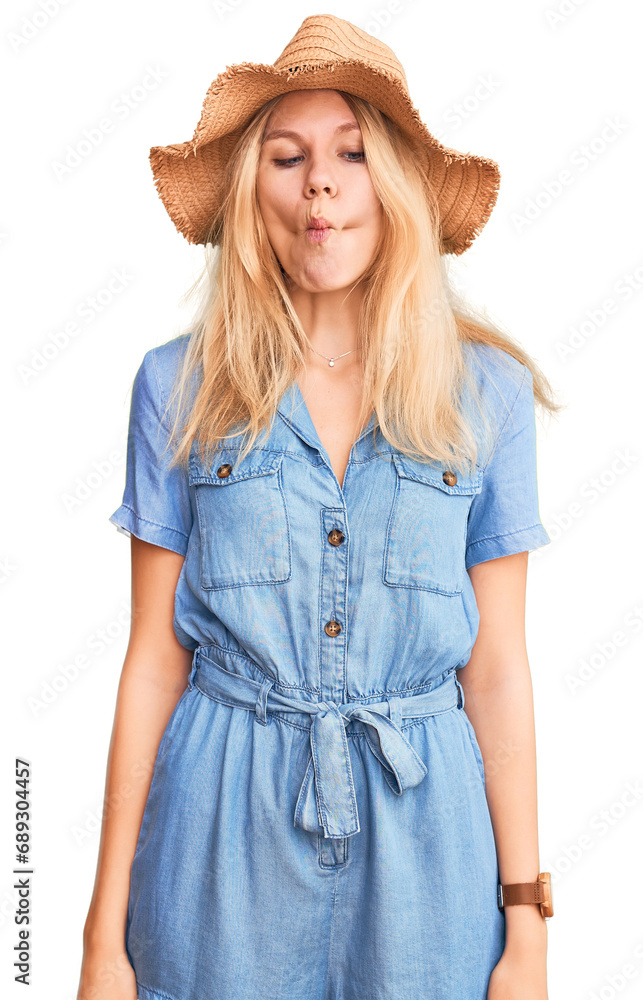 Fototapeta premium Young beautiful blonde woman wearing summer hat and dress making fish face with lips, crazy and comical gesture. funny expression.