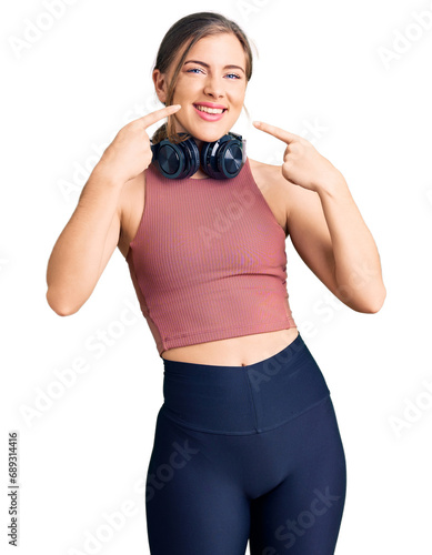 Beautiful caucasian young woman wearing gym clothes and using headphones smiling cheerful showing and pointing with fingers teeth and mouth. dental health concept.