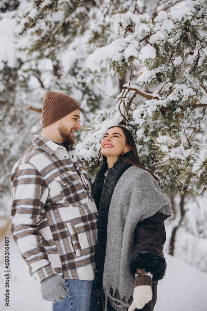 A young happy and loving couple is standing under a pine tree from which snow falls in the mountains in winter.