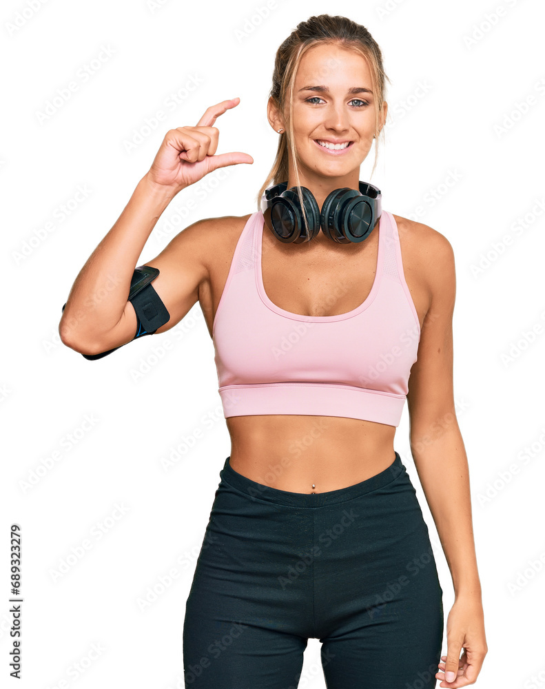 Young blonde woman wearing gym clothes and using headphones smiling and confident gesturing with hand doing small size sign with fingers looking and the camera. measure concept. Young blonde woman wearing gym clothes and using headphones smiling and confident gesturing with hand doing small size sign with fingers looking and the camera. measure concept.