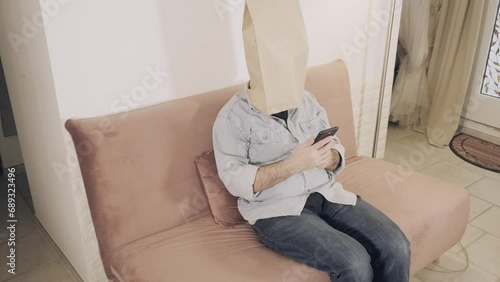 A man wearing a breadbag face, sitting on the sofa at home, no expression on his paper mask; checking his smartphone, bored, throwing it away.
 photo