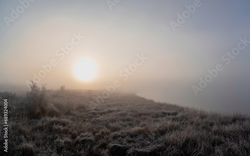The sun rises over the field in a thick fog. Minimalistic natural foggy landscape. The shore of the lake is barely visible in the morning fog. Panoramic view.