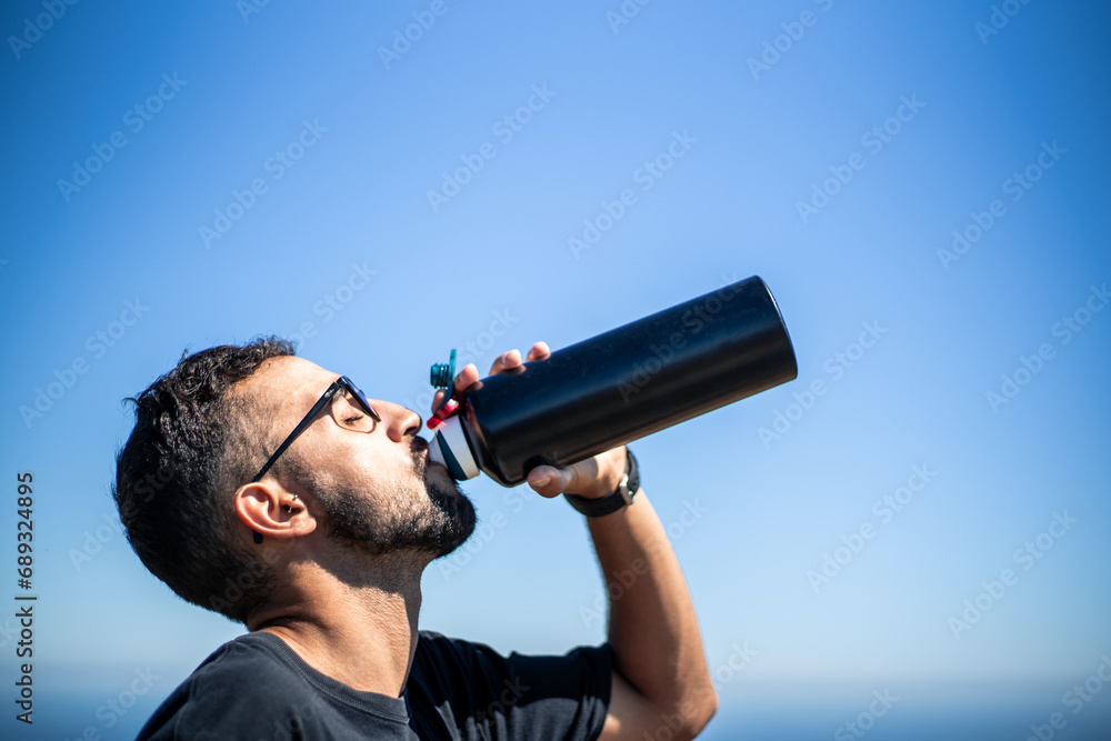 Young Caucasian man of North African nationality thirstily drinking water from a black unbranded canteen or bottle on a very sunny and hot summer day. Profile view.