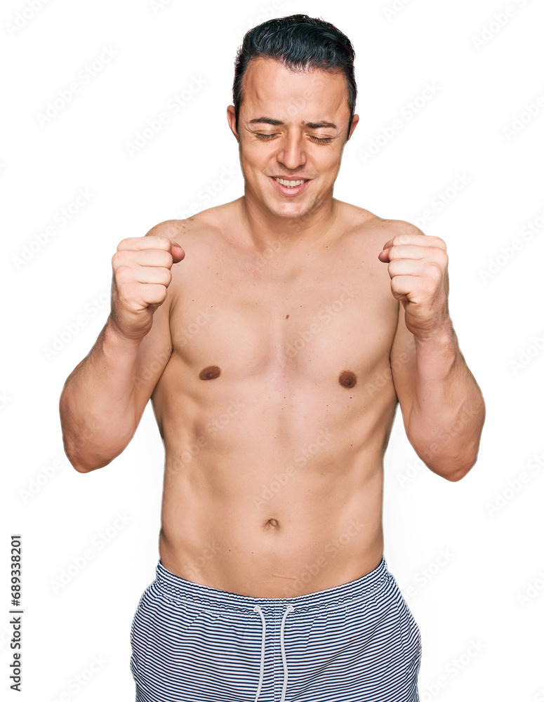 Handsome young man wearing swimwear shirtless excited for success with arms raised and eyes closed celebrating victory smiling. winner concept. Handsome young man wearing swimwear shirtless excited for success with arms raised and eyes closed celebrating victory smiling. winner concept.