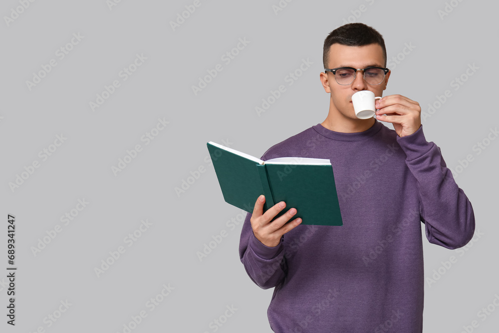 Handsome young man reading book and drinking coffee on grey background Handsome young man reading book and drinking coffee on grey background