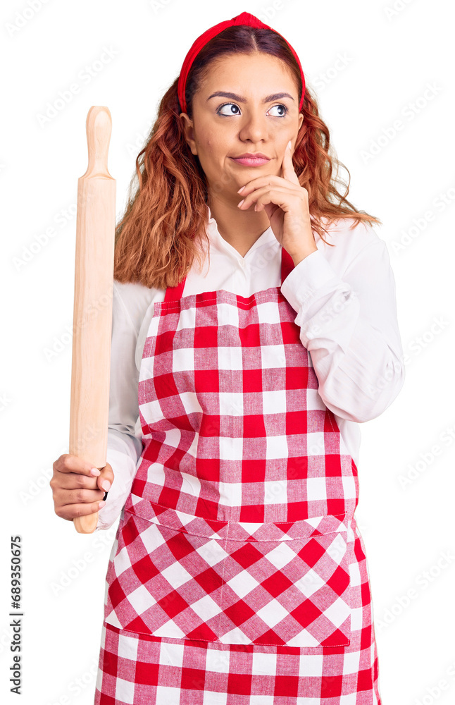 Young latin woman wearing apron holding kneader serious face thinking about question with hand on chin, thoughtful about confusing idea Young latin woman wearing apron holding kneader serious face thinking about question with hand on chin, thoughtful about confusing idea
