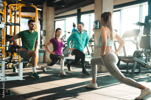 Group of athletic people exercising lunges in a health club. Team of happy gym newcomers following instructions of their female fitness instructor. Female coach performing exercises with a group.