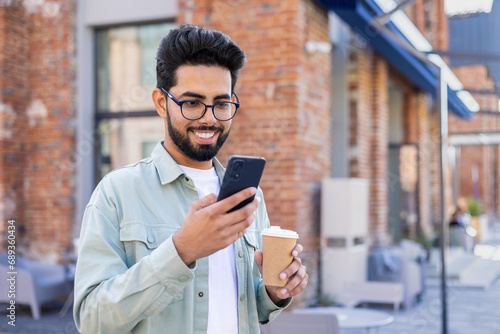 A happy Indian man is standing on the street, holding a cup of coffee, smiling and looking at the smartphone screen.
