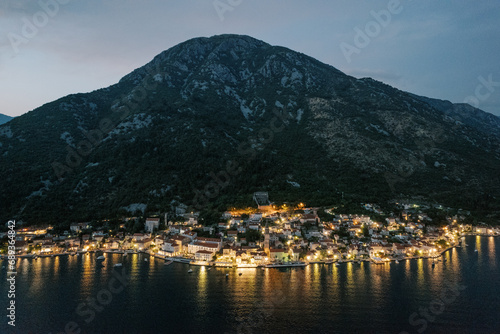 Coast of Perast glowing with night lights at the foot of the mountains. Montenegro. Aerial view