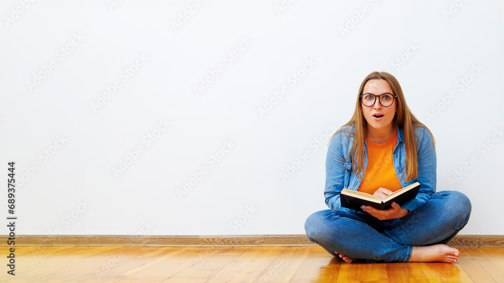 Surprised woman with glasses sits on the floor against white wall holding book Surprised woman with glasses sits on the floor against white wall holding book