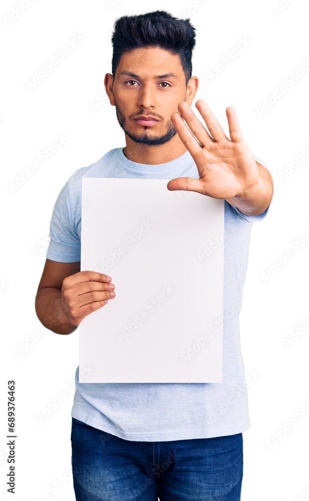 Handsome latin american young man holding cardboard banner with blank space with open hand doing stop sign with serious and confident expression, defense gesture Handsome latin american young man holding cardboard banner with blank space with open hand doing stop sign with serious and confident expression, defense gesture
